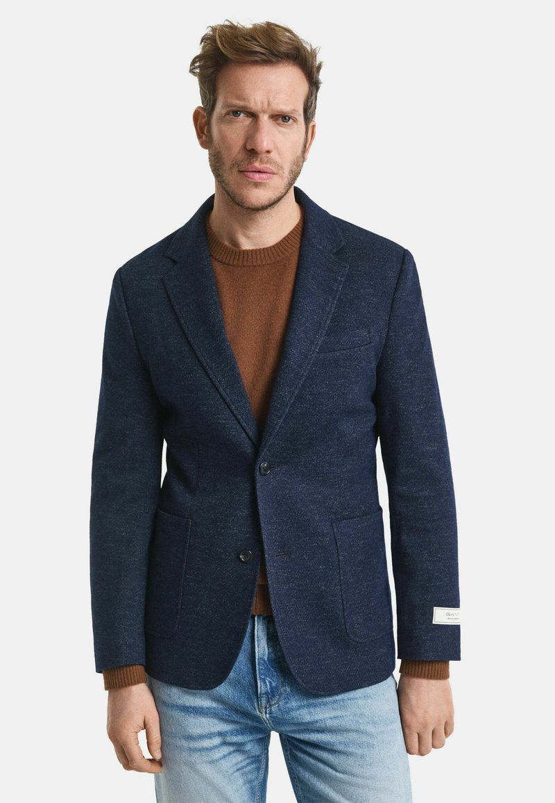 Marineblauer Woll-Blazer mit Revers, zwei Vordertaschen und sichtbarem Etikett. Trägt man über einem braunen Pullover und kombiniert mit hellen Jeans.