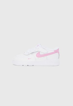 Biały sneaker Nike z różowymi logo w kształcie znaku Swoosh, perforowanym noskiem i paskiem na rzep na jednolitym jasnoszarym tle, widok z boku.