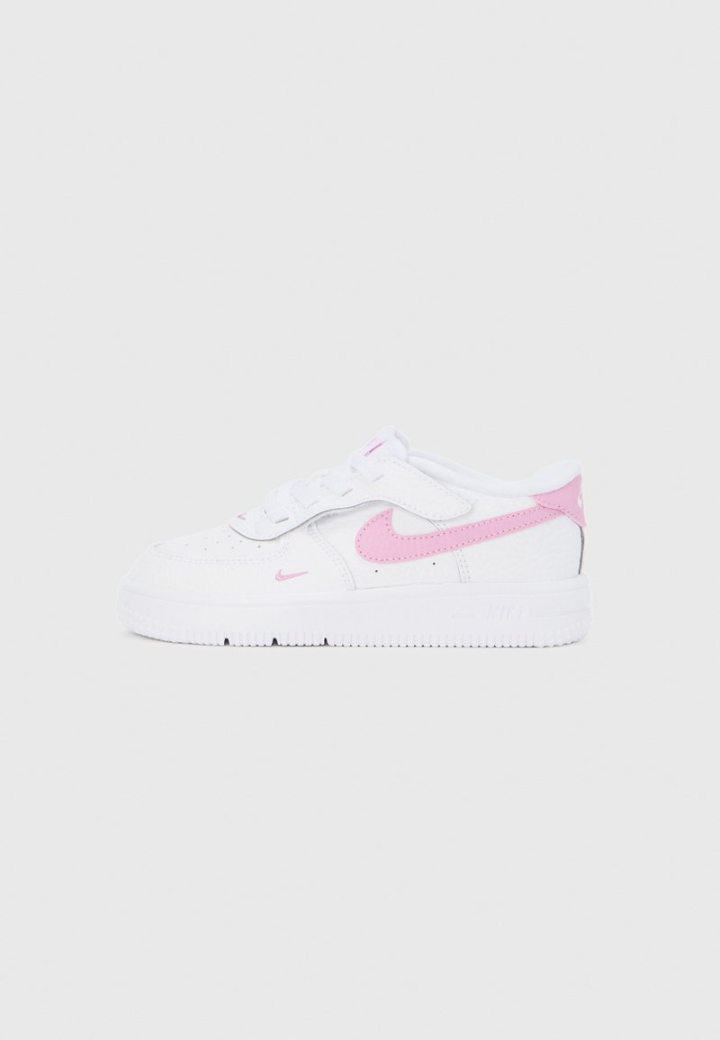 Λευκό αθλητικό παπούτσι Nike με ροζ λογότυπα σβούρας, διάτρητη μύτη και λουράκι με βέλκρο σε μονόχρωμο απαλό γκρι φόντο, πλάγια όψη.