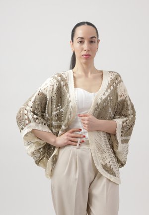 Vrouw met donker haar naar achteren gebonden, draagt een beige en crème gehaakte sjaal over een witte top en lichtbeige broek, staand tegen een effen achtergrond.