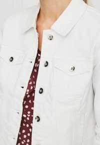 Vit denimjacka med silverknappar som bärs över en vinröd topp med ljusa, oregelbundna fläckar.