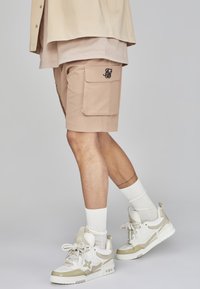 Pantaloni cargo beige con tasche laterali e logo ricamato, abbinati a sneaker bianche e beige e calzini bianchi a costine.