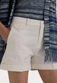 White shorts with a tailored design, featuring a front pocket with a button and a rolled hem. Paired with a blue-striped woven scarf.