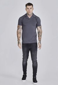 Szürke, rövid ujjú, testhezálló poloing, sötét farmernadrággal és fekete tornacipővel viselve, karokon tetoválásokkal.