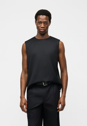 Homme portant un haut noir sans manches et un pantalon noir avec ceinture, debout devant un fond gris clair uni, faisant face à l'avant.