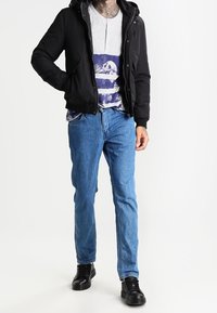 Homme portant une veste noire, un t-shirt blanc avec un motif de crâne, un jean bleu et des baskets noires, debout sur un fond blanc.