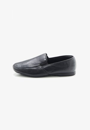 Zapato negro de piel sin cordones con una textura suave, punta redondeada y diseño minimalista; presenta costuras sutiles y una suela de goma plana.