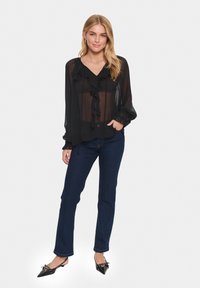 Blusa nera trasparente con fronte arricciato e maniche lunghe, abbinata a jeans in denim scuro e scarpe nere a punta. Design semplice e tessuto leggero.