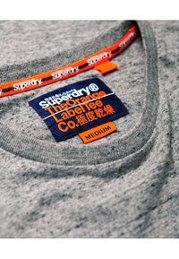 Grå Superdry T-shirt med en orange och marinblå etikett som säger "The Orange Label Tee Co." och storleksmärkning medium.
