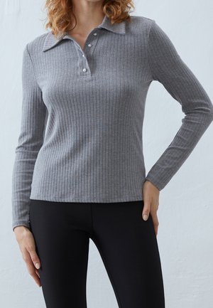 Mujer que lleva un jersey tipo polo gris de manga larga acanalado con botones de perla y pantalones ajustados negros sobre un fondo claro y liso.