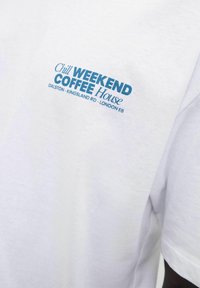 Balta medvilninė marškinėlių su mėlynu tekstu, kuriame rašoma "Chill WEEKEND Coffee House", o kairėje krūtinės pusėje pateikti užrašai su lokacija.
