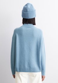 Persoană purtând o căciulă tricotată de un bleu deschis și un pulover cu model ribat, stând cu fața către un perete alb, cu pantaloni albi vizibili din spate.