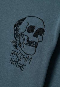Katoenen T-shirt in teal met zwart geborduurde schedelafbeelding. Onder de schedel staat de tekst "RHYTHM OF NATURE". Zachte textuur, casual ontwerp.