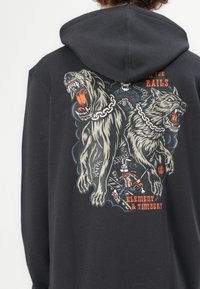 Sudadera gris oscuro con un gráfico que presenta dos perros feroces, cadenas y un personaje en las vías del tren. Tejido suave con un corte relajado.