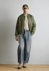 Olivenfarget bomberjakke, beige strikket topp, løse lyseblå jeans og brune ankelstøvler. Fullført med en grønn caps.