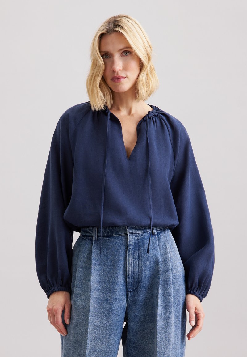 Blusa blu navy a maniche lunghe con scollatura a V e collo arricciato. Indossata con jeans in denim azzurro a vita alta con righe verticali.
