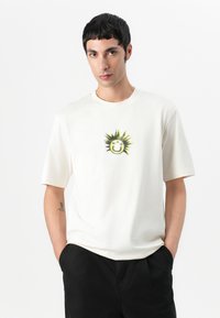 T-shirt en coton couleur crème à manches courtes, présentant un graphique de soleil en bande dessinée avec un visage souriant et des rayons en pointes verts et noirs.