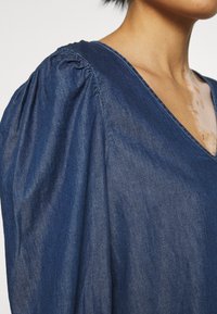 La blusa in denim presenta un profondo scollo a V, maniche svasate e una texture liscia di un blu scuro. Le cuciture visibili aggiungono dettagli al design.