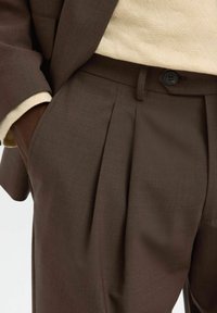Brązowe spodnie w kant wykonane z gładkiego materiału, z zapięciem na guzik, bocznymi kieszeniami i dopasowanym krojem.