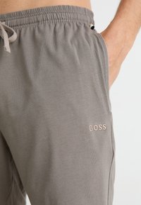 Grå bomullsjoggesko med elastisk midjebånd, snor og sidelommer. Har et lite rosa brodert "BOSS"-logo.