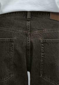 Gros plan de l’arrière d’un jean en denim gris foncé avec coutures visibles, passant de ceinture et deux poches arrière carrées, porté avec une chemise blanche.