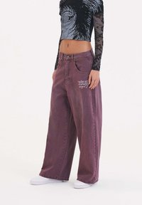 Breda ben, högmidjade lila denimbyxor med framfickor, med broderad vit text på vänster höft. Slät struktur, avslappnad design.