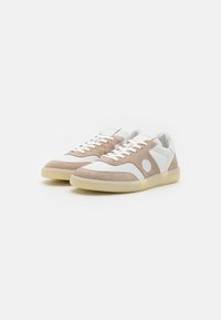 Brown suede and white leather sneaker with a rounded toe, flat laces, and a beige rubber sole. Features a circular logo on the side.
