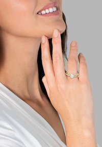 Bague en or avec une grande pierre précieuse claire et un anneau texturé, portée sur une main touchant le menton, sur un fond neutre.