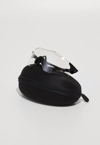 Przezroczyste sportowe okulary przeciwsłoneczne leżą na czarnym, owalnym etui twardym z zamkiem błyskawicznym. Okulary mają smukły design z minimalistyczną ramką.