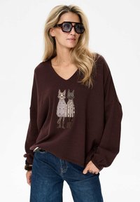 Brązowy, oversize sweter z dekoltem w serek, z haftowanym wzorem przedstawiającym dwa kreskówkowe koty ubrane w pasiaki i wzorzyste stroje. Ciemne dżinsy.