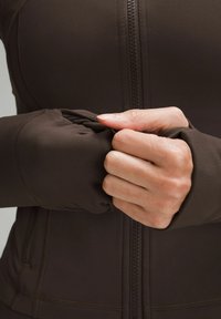 Brun zip-up jakke med tætsiddende manchetter, lavet af strækbart materiale. Detaljer inkluderer synlige syninger og et nærbillede af hænder, der holder fast i stoffet.