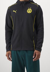 Uomo che indossa una felpa nera con zip e logo Puma giallo, insieme all'emblema BVB 09, abbinata a pantaloni verde scuro, in piedi su uno sfondo bianco.