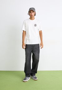 Hvit kortermet t-skjorte med et rundt grafisk trykk på brystet, mørkegrå jeans og grå joggesko med hvite detaljer, stående på grønt gulv.