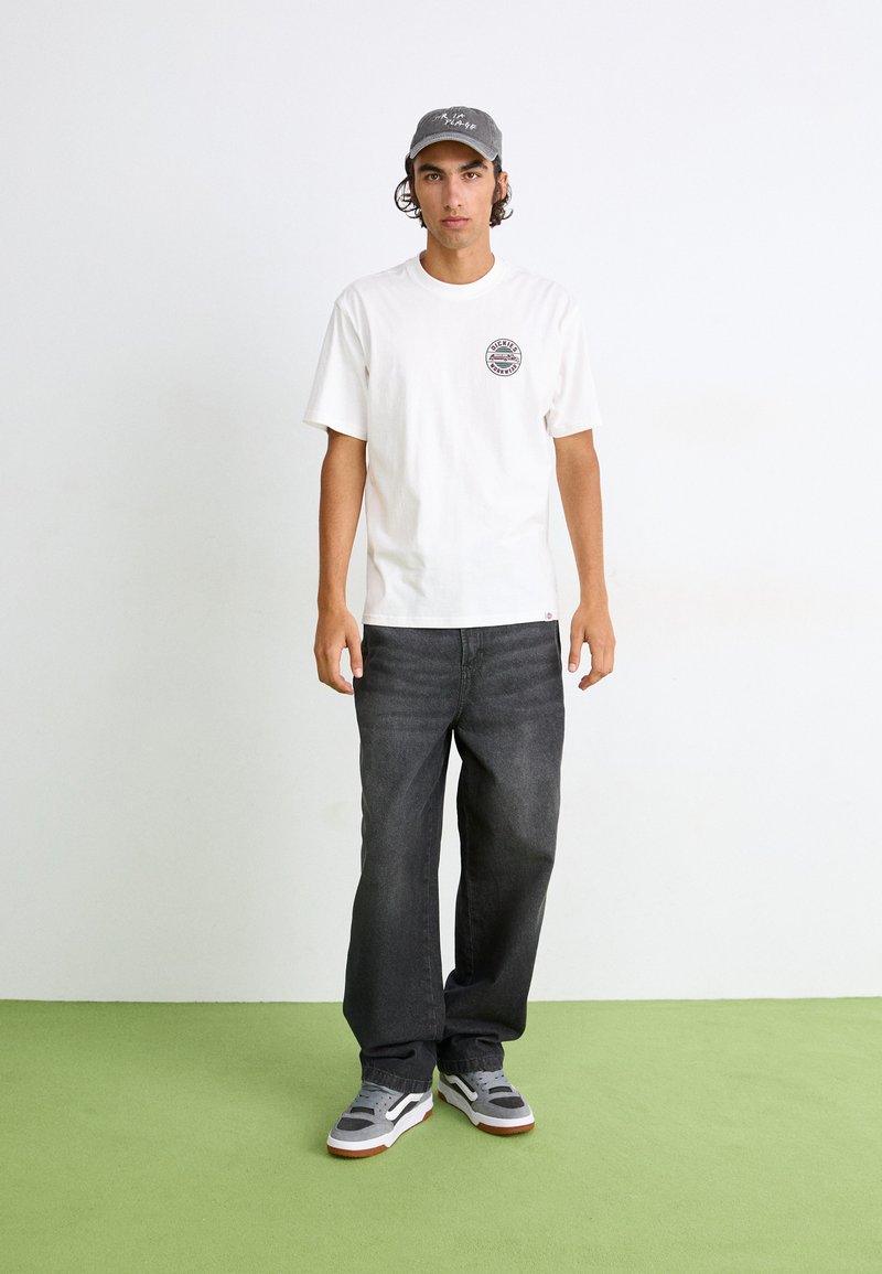 Weißes T-Shirt mit kurzen Ärmeln und runder Grafik auf der Brust, dunkelgraue Jeans und graue Sneaker mit weißen Akzenten, stehend auf grünem Boden.