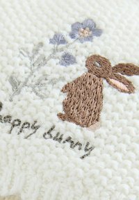Strickstoff in Creme zeigt einen bestickten braunen Hasen mit gestickten Blumen und dem Text "glücklicher Hase" in grauem Faden, der Textur und Details präsentiert.