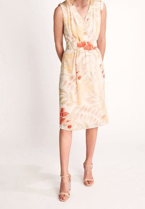 Femme portant une robe beige sans manches avec des motifs floraux orange et jaune, associée à des sandales beiges à talons hauts, debout devant un fond blanc.