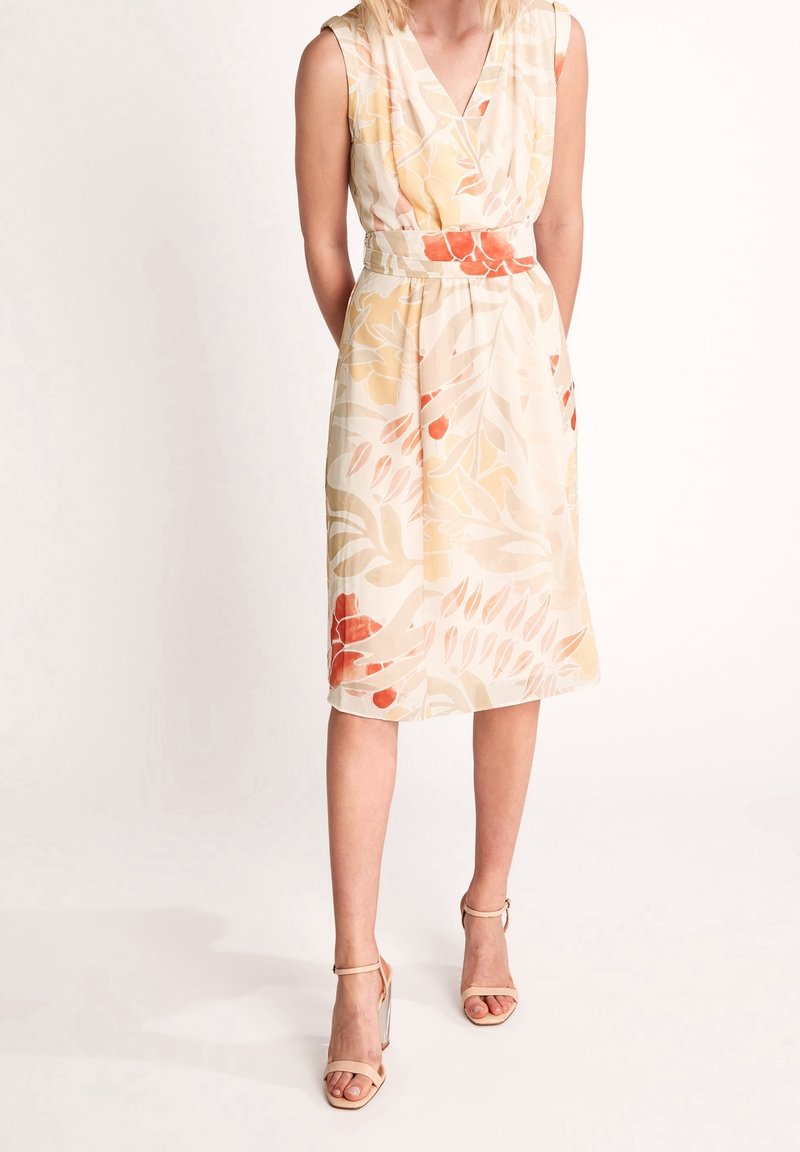 Femme portant une robe beige sans manches avec des motifs floraux orange et jaune, associée à des sandales beiges à talons hauts, debout devant un fond blanc.