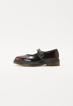 Chaussure Mary Jane noire brillante avec une semelle en caoutchouc translucide marron et une bride à boucle dorée sur le dessus du pied sur fond blanc.