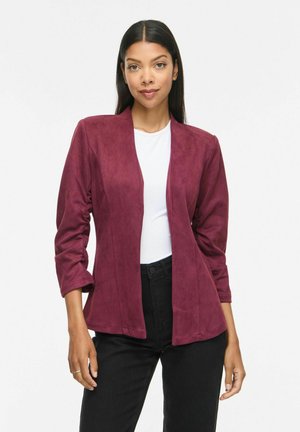 Femme aux longs cheveux foncés portant un blazer bordeaux ouvert, un haut blanc et un pantalon noir, debout devant un fond blanc uni.