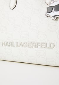 White leather handbag with embossed faces pattern and "KARL LAGERFELD" logo. Features a playful cartoon charm with sunglasses.
