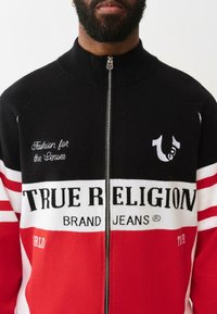 Pulover cu fermoar în negru, roșu și alb. Prezintă un branding îndrăzneț "TRUE RELIGION", modele cu dungi și broderie cu mesajul "Fashion for the Senses".