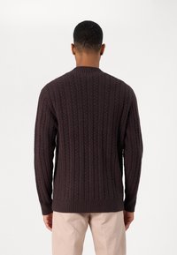 Pulover gros cu tricotaj cu model de cablu, de culoare maro închis, cu guler rotund, mâneci lungi și mansete și tiv cu nervuri. Textura este groasă și texturată.
