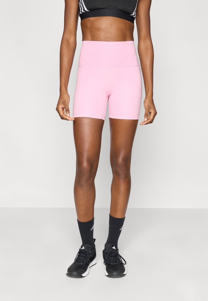 Ροζ αθλητικά σορτς με ψηλή μέση και πλαϊνές ραφές, κατασκευασμένα από λείο ύφασμα. Φοριούνται με μαύρα αθλητικά παπούτσια και κάλτσες για ολοκληρωμένη εμφάνιση.