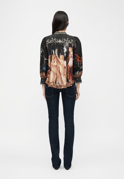 Femme aux longs cheveux bruns portant un chemisier noir orné d'une œuvre d'art de style Renaissance dans le dos, associée à un jean foncé ajusté, debout de dos.