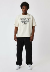 Camiseta gráfica beige con el estampado "Project Paris" y pantalones cargo negros; cuenta con bolsillos y zapatillas blancas, todo sobre un fondo neutro.