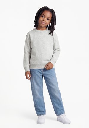 Junges Mädchen mit geflochtenem Haar steht da und trägt einen hellgrauen Pullover, blaue Jeans und weiße Sneakers vor weißem Hintergrund.