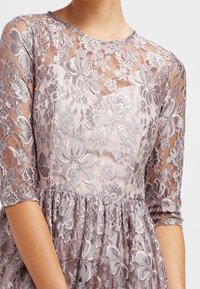 Spetsklänning med blommönster i mjuk mauve och silver, med en figurnära överdel och en full, rynkad kjol. Genomskinliga långärmar.