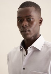 Lichtgrijs T-shirt met korte mouwen en een button-down kraag, met een patroon in marineblauwe accenten. Heeft een borstzak en een subtiele textuur.