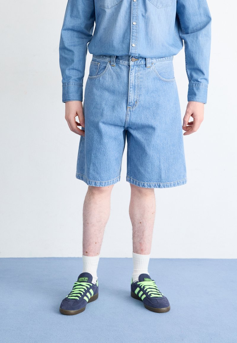 Lyseblå denim shorts med lige snit, prydet med forlommer. Kombinationen inkluderer en matchende lyseblå skjorte med knapper og sorte sneakers.