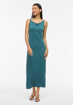 Naine kandmas türkiissinist varrukateta maksikleiti volditud dekolteega ja musti rihmikveri sandaale, seistes ja naeratades valge tausta ees.